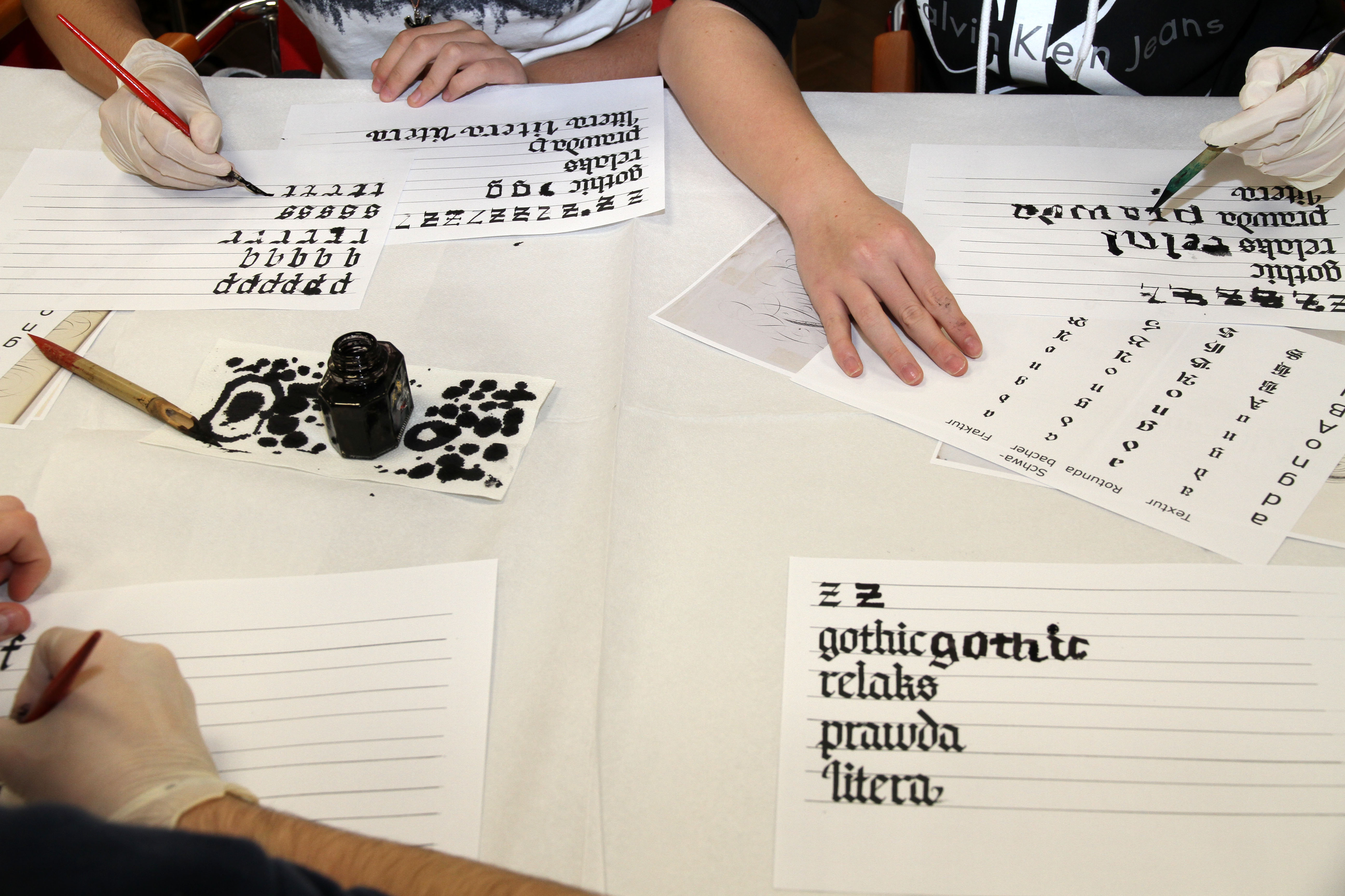 Uczestnicy warsztatów kaligrafują litery na kartkach papieru. 
