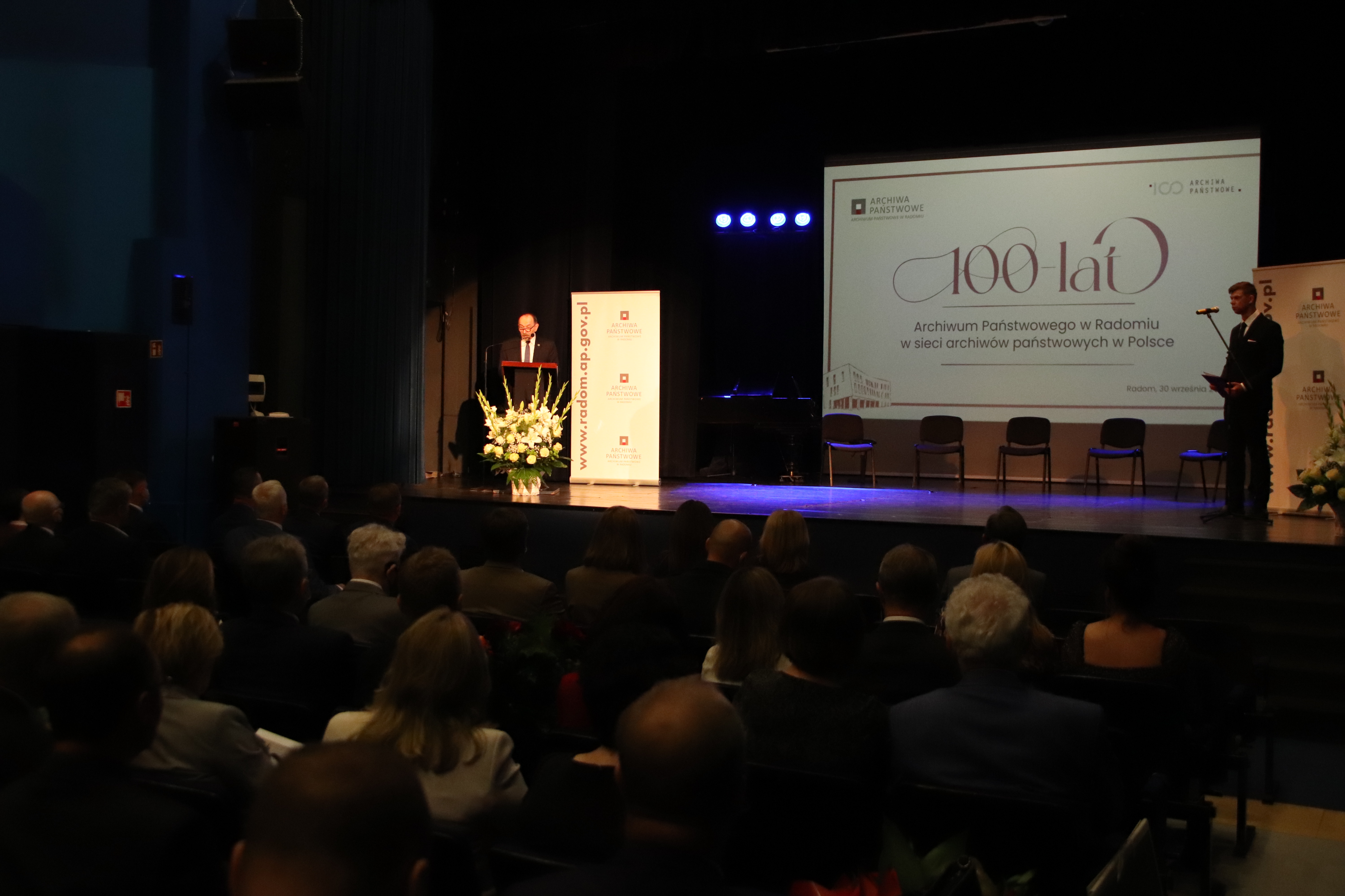 Dyrektor Jaroszek przemawia na scenie, oświetlony reflektorem, za nim slajd prezentacji z napisem 100 lat Archiwum Państwowego w Radomiu w sieci archiwów państwowych w Polsce.