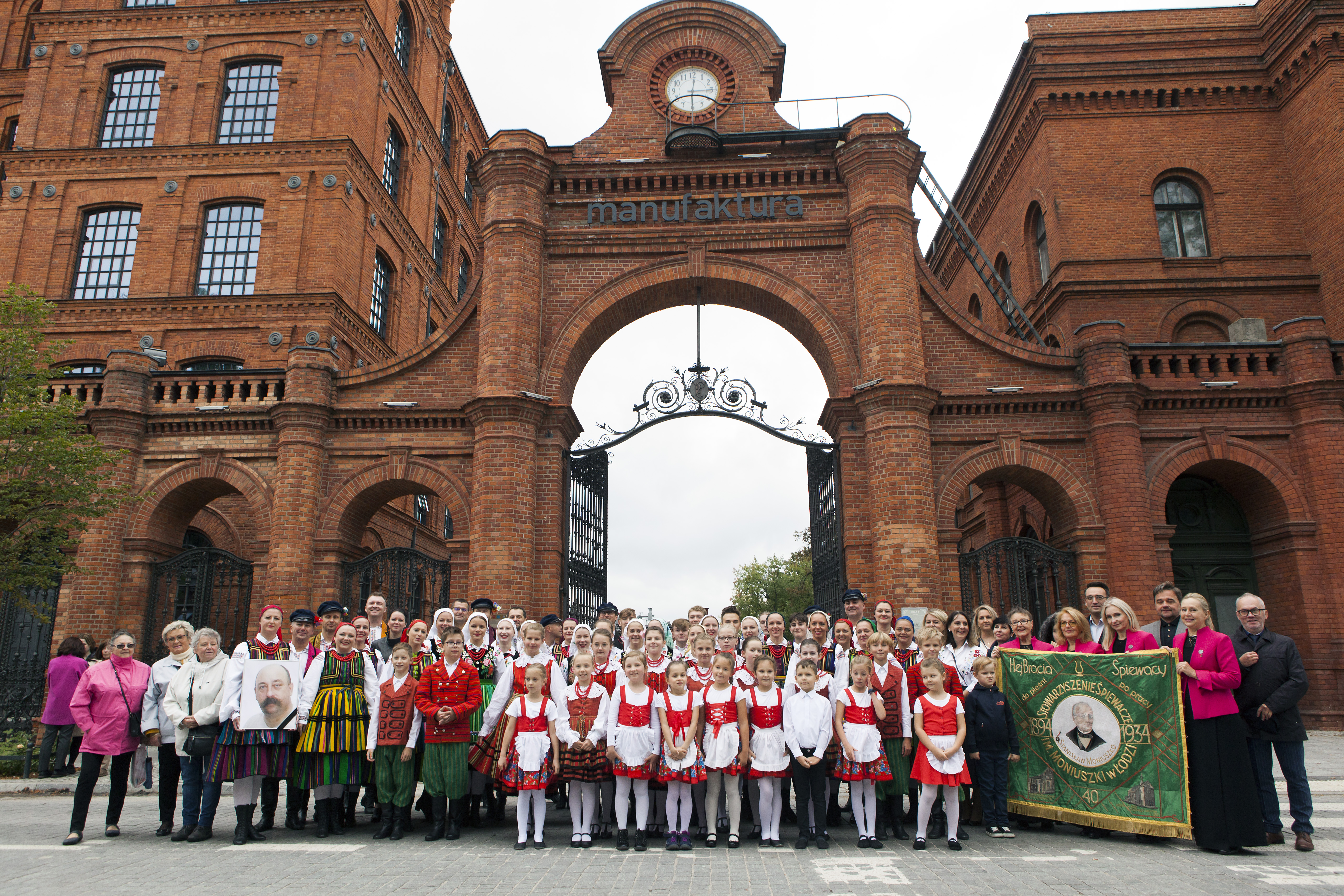 Zdjęcie grupowe gości wydarzenia przed bramą Manufaktury. 
