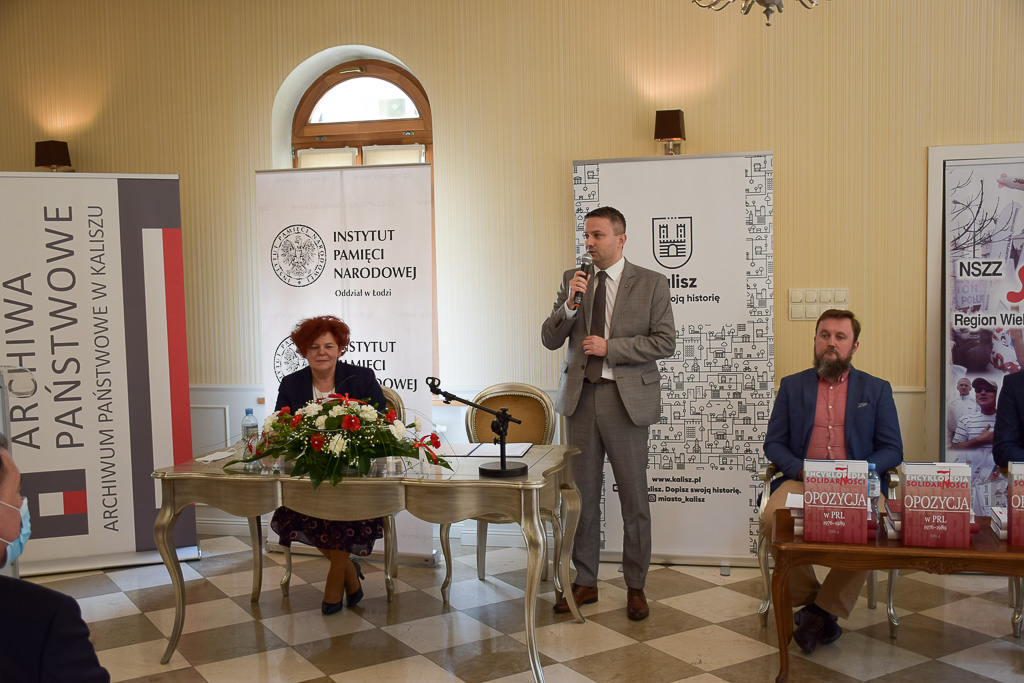 Dr Sebastian Pilarski przemawia, dr Grażyna Schlender i dr Grzegorz Wołk siedzą przy dwóch oddzielnych stołach. 