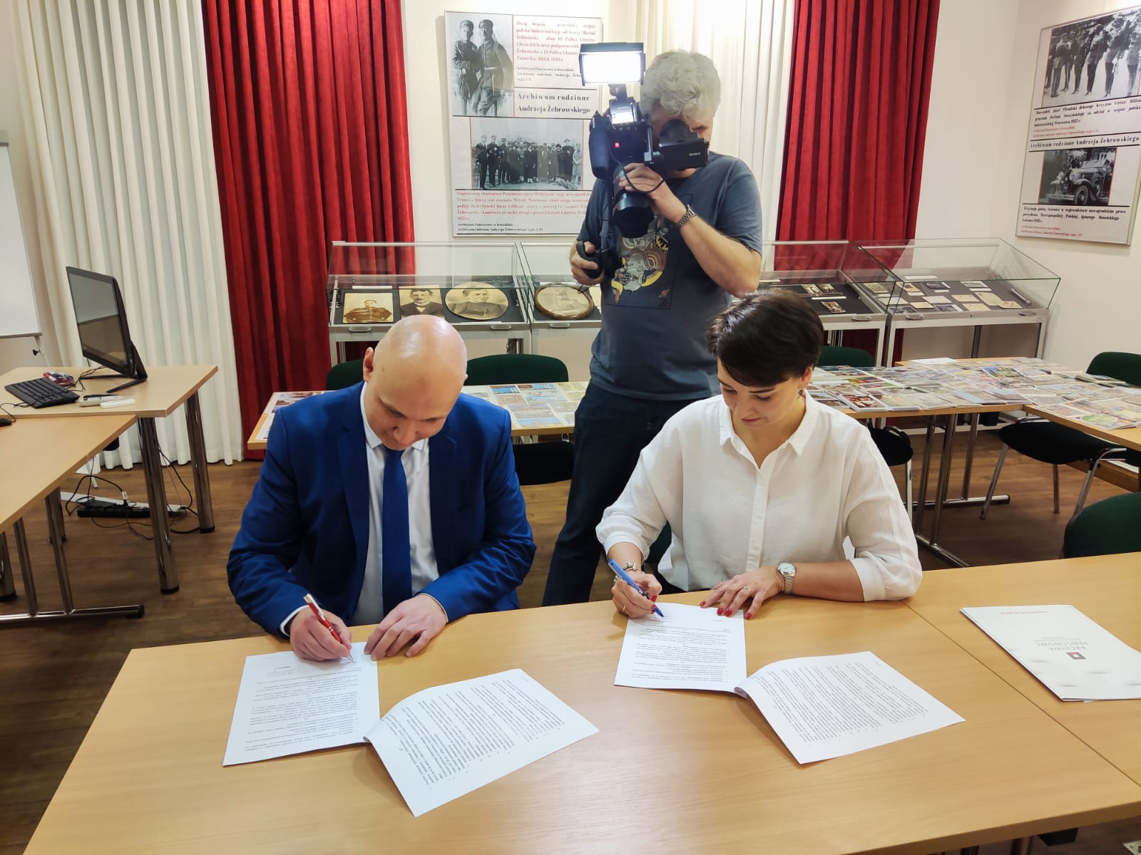 Wojciech Stylski i dyrektor Królczyk podpisują umowę. Za nimi mężczyzna z kamerą. Z tyłu stół zapełniony pocztówkami.