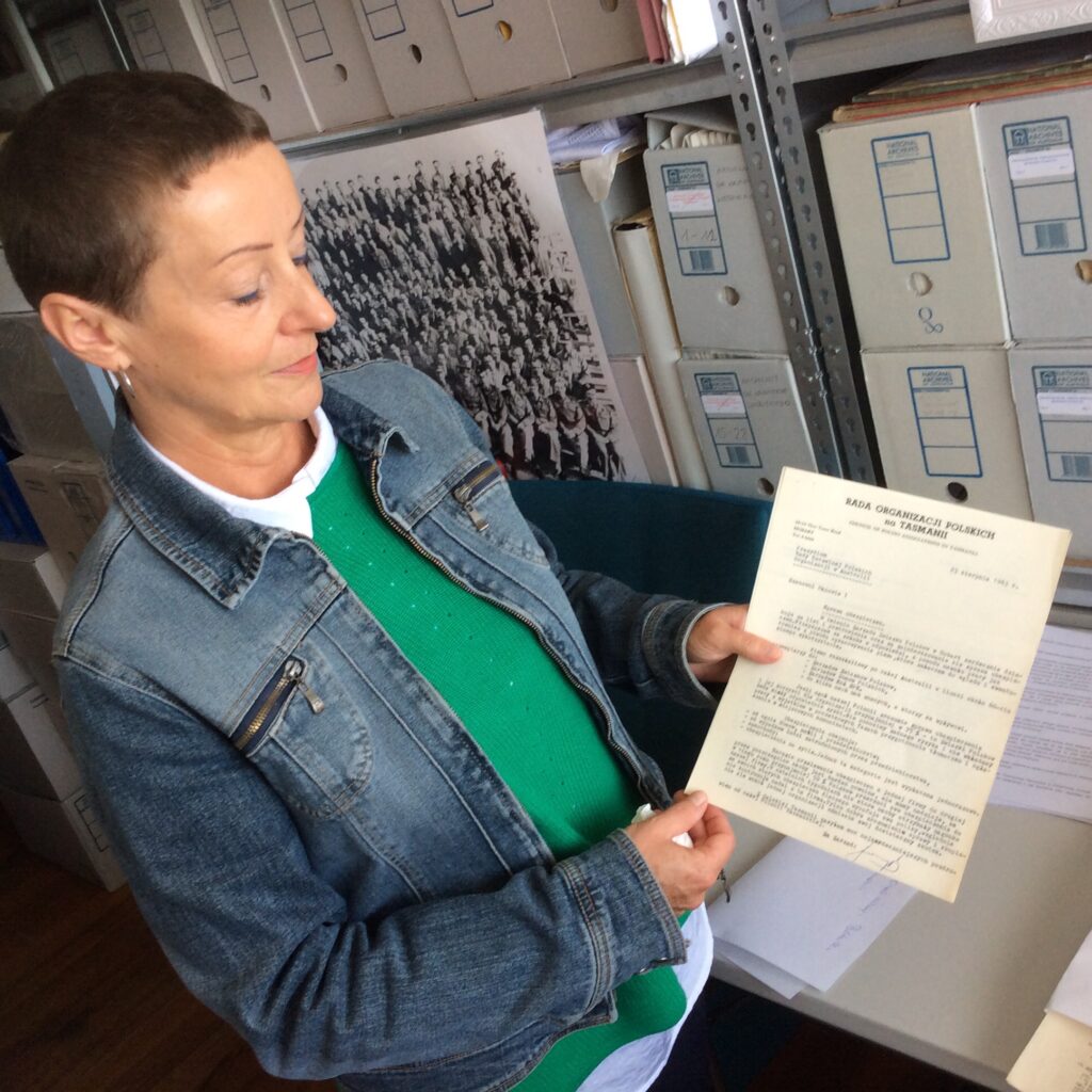 Barbara Grzybek trzyma w dłoniach dokument i pokazuje go do aparatu. W tle regału z pudłami archiwalnymi w których przechowywane są dokumenty.