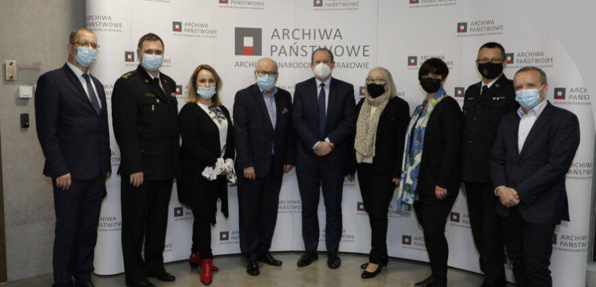 Dziewięć dorosłych osób w maskach na twarzach i formalnych strojach stoi w rzędzie do zdjęcia. Za nimi znajdują się banery Archiwa Państwowe i logo organizacji wskazujące na oficjalne wydarzenie lub spotkanie w Archiwum Państwowym.