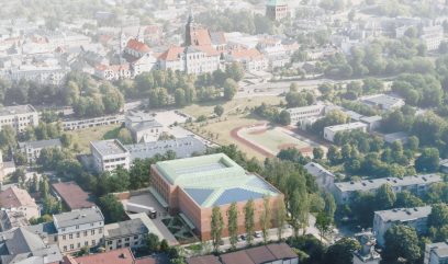 Widok z lotu ptaka na miasto z nowoczesnym, prostokątnym budynkiem z cegły ze szklanym dachem na pierwszym planie, otoczonym drzewami. Obok znajduje się boisko sportowe z bieżnią. W tle widoczne są zabytkowe budynki i kościół z wysoką wieżą.