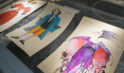 Trzy kolorowe rysunki kostiumów w szklanej gablocie.
