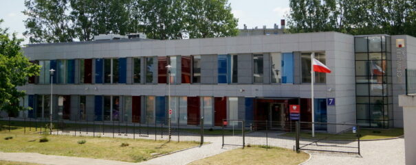 Budynek archiwum z dużą ilością przeszkleń zabarwionych na czerwono i niebiesko. Przed budynkiem trawnik oraz ogrodzenie. Na maszcie przed budynkiem powiewa polska flaga.