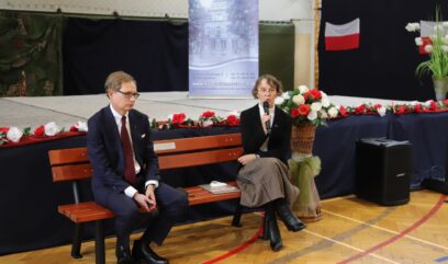 Jolanta Szyłkowska i Krzysztof Kowalczyk siedzą na ławce na środku sali. Jolanta Szyłkowska mówi do mikrofonu, który trzeba w dłoni.