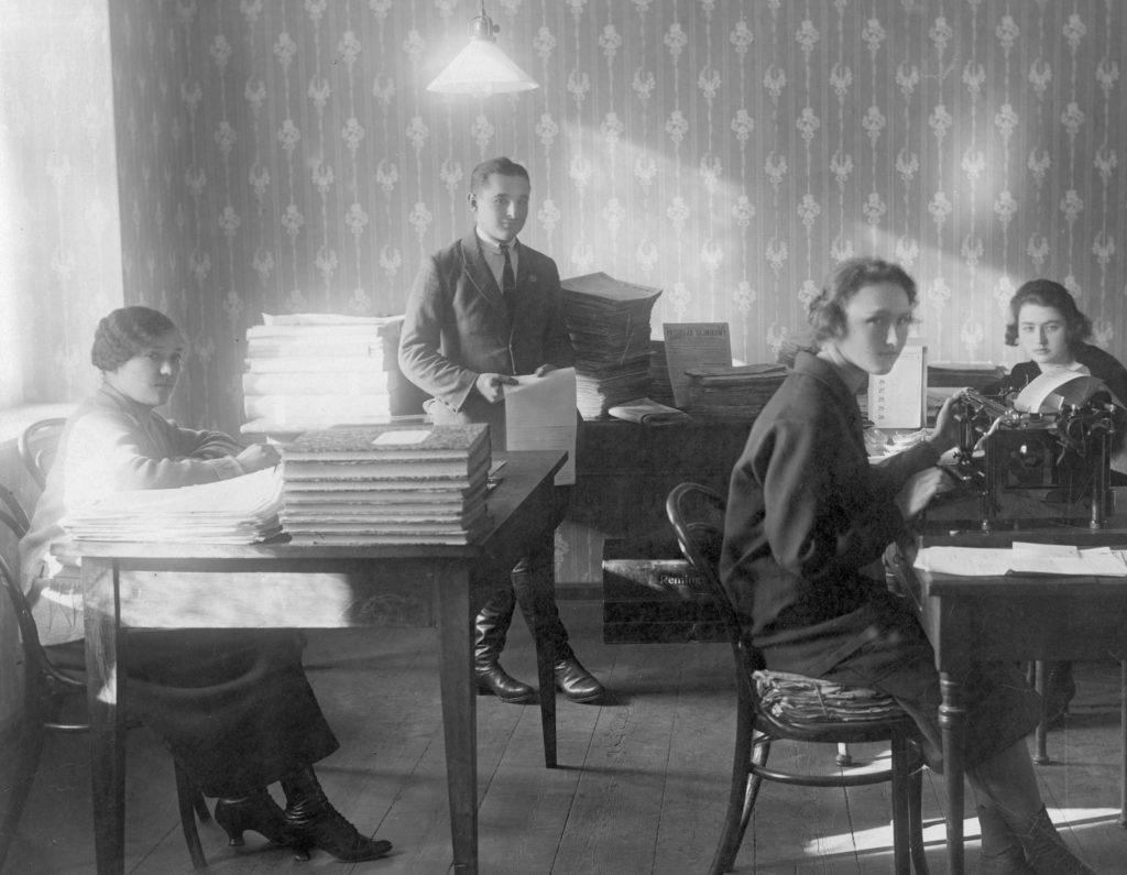 Pomieszczenie biurowe. Trzy kobiety przy pracy siedzą przy biurkach. Na biurkach urządzenia biurowe oraz księgi i dokumenty. Na dalszym planie stoi mężczyzna. W tle stos „Przeglądów Sejmikowych” przeznaczonych do wysyłki.