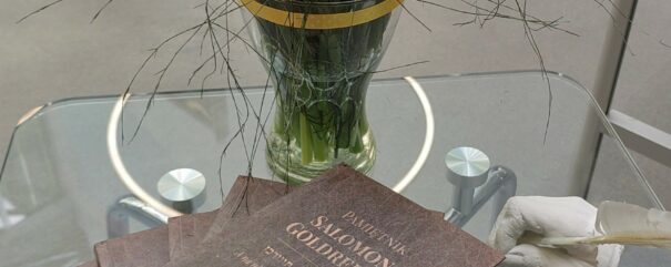 Kilka egzemplarzy książki leży jedna na drugiej na szklanym stoliku. Obok stoi wazon z żonkilami.