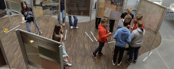 13 uczniów spaceruje po sali wystawowej między planszami wystawy i oglądają je. Na zdjęciu widoczne 6 plansz wystawy, za nimi pod ścianami ustawione szklane gabloty z oryginalnymi archiwaliami.