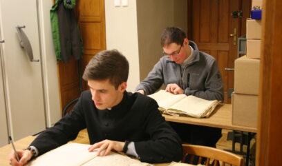 Dwóch wolontariuszy siedzi przy stoliku i numeruje ołówkiem kolejne strony księgi.