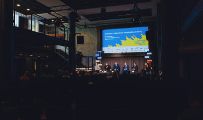6 mężczyzn siedzi na scenie przy stole konferencyjnym. W tle niebiesko-żółty baner informacyjny konferencji.