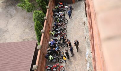 Co najmniej 12 motocykli stoi w rządku na dziedzińcu budynku archiwum. Dookoła spacerują ludzie, oglądający motocykle.