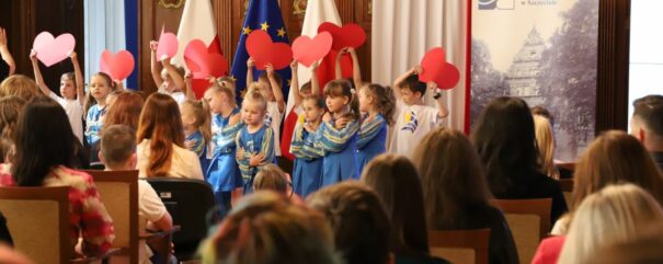 Na scenie, na tle flagi Polski i Unii Europejskiej, występują dzieci w wieku przedszkolnym. W dłoniach mają uniesione do góry papierowe serca. Fot. Daniel Wróbel (Kuratorium Oświaty w Szczecinie)