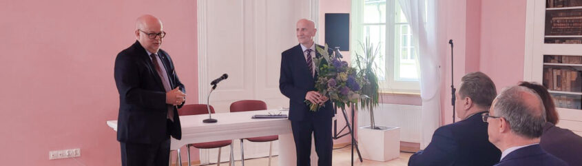 Dwóch, ubranych w garnitury mężczyzn stoi na środku sali – jeden przemawia, drugi w ręku trzyma bukiet kwiatów. Przed nimi w dwóch rzędach siedzą elegancko ubrani ludzie.
