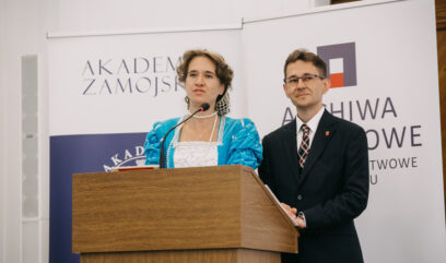 Dyrektor Żygawski i dyrektor Polańska stoją za mównicą. Kobieta przebrana jest w niebieską suknię z epoki.