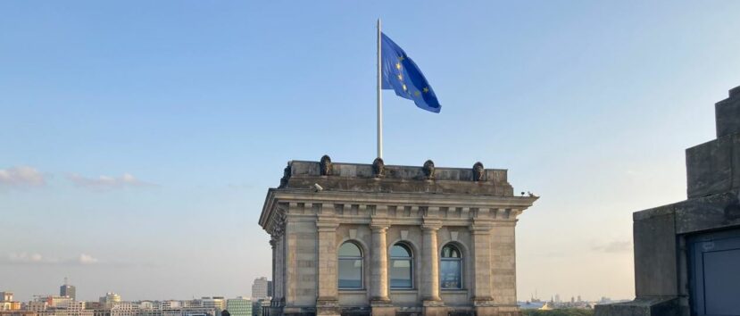 Dach budynku ozdobiony rzeźbami. Na wieży dachu powiewa na wietrze flaga Unii Europejskiej. W tle panorama miasta.