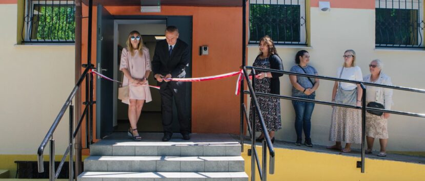 Budynek, do którego prowadzą schody. Po obu stronach schodów barierka. Na najwyższym schodku stoi kobieta w różowej sukience i mężczyzna w garniturze. Mężczyzna przecina biało-czerwoną wstęgę przywiązaną po obu stronach do barierki.