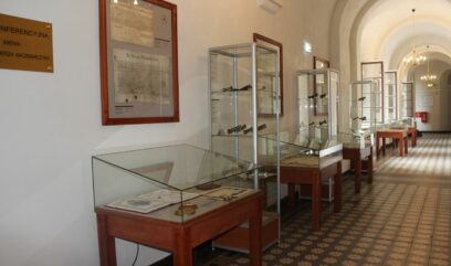 Wzdłuż korytarza stoją przeszkolone gabloty z eksponatami z wystawy – pięć niższych i trzy wyższe.