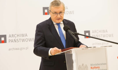 Premier Gliński przemawia do mikrofonu. Za nim ścianka z logo Archiwa Państwowe i ekran z logo MKiDN.