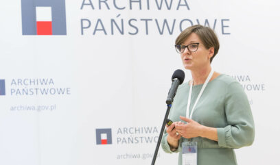 Katarzyna Kiliszek przemawia do mikrofonu. Za nią ścianka z logo Archiwa Państwowe.