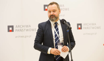 Dyrektor Tomasz Makowski przemawia do mikrofonu. Za nim ścianka z logo Archiwa Państwowe.