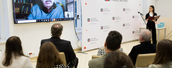Na dużym ekranie Czesław Czapliński w łączeniu na żywo. Obok stoi konferansjerka. Przed nimi siedzi publiczność.