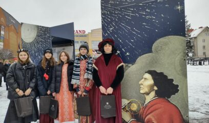 Na ziemi leży śnieg. Ubrani w historyczne stroje z epoki Mikołaja Kopernika chłopak i cztery dziewczyny stoją przed wystawą.