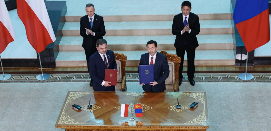 4 mężczyzn stoi, z tyły przy schodach prezydenci Andrzej Duda i Ukhnaagiin Khürelsükh, przy stole dyrektorzy archiwów Paweł Pietrzyk i Enkhbaatar Samdan. Dyrektorzy trzymają w rękach teczki z memorandum.