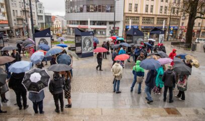 Plac miejski. Pada deszcz. Na placu przed wystawą stoi tłum ludzi z parasolkami.