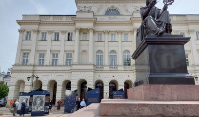 Pomnik Mikołaja Kopernika w Warszawie. Za nim stoją bryły wystawy. Obok wystawy przechadzają się ludzie.