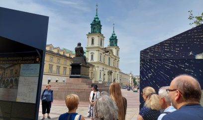 7 osób ogląda plansze wystawy. W tle pomnik Mikołaja Kopernika w Warszawie i kościół Świętego Krzyża.