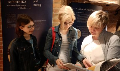 Podziemia. Trzy kobiety przeglądają książkę. Na ścianie wisi plansza wystawy „Ślady Mikołaja Kopernika w Archiwum Państwowym w Gdańsku”.