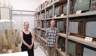 Magazyn archiwalny. Na półkach stoi kilkadziesiąt telewizorów z różnych lat. Obok półek stoją mężczyzna i kobieta.
