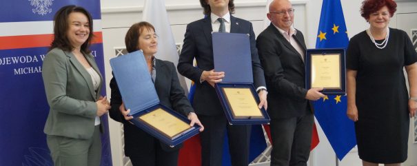 4 osoby stoją na podeście. Trzech laureatów trzyma dyplomy w eleganckich pudłach.