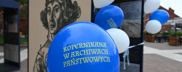 Niebieski balonik z napisem: „Kopernikana w Archiwach Państwowych”. W tle granatowa plansza wystawy ze zdjęciem mężczyzny.