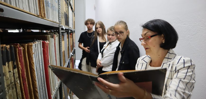Grupa uczniów w magazynie archiwalnym. Archiwistka pokazuje im dokument.