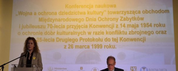 Dyrektor Wiercińska stoi za pulpitem. Obok za stołem siedzi prezes Polskiego Komitetu Błękitnej Tarczy Krzysztof Sałaciński.