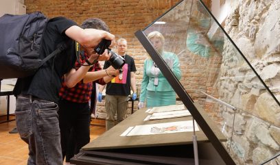 Sala wystawowa. 2 osoby robią zdjęcia dokumentu wyłożonego w szklanej gablocie. Obok stoi archiwistka i archiwista.