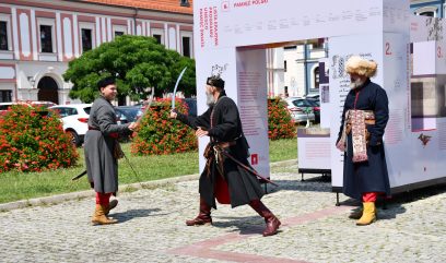 Dwie osoby w strojach szlachciców walczą na szable. Przygląda się temu trzeci mężczyzna przebraniu szlachcica.