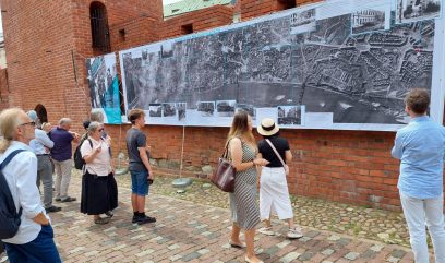 Grupa ludzi ogląda wystawę wywieszoną na murach Barbakanu.