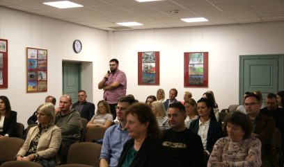 Kilkadziesiąt osób siedzi na Sali konferencyjnej. Jeden z uczestników stoi i mówi do mikrofonu.