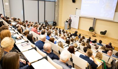 Aula wykładowa. Dyrektor Pietrzyk przemawia do mikrofonu. Za nim rollupy z logo Archiwa Państwowe. Przed nim siedzi publiczność.