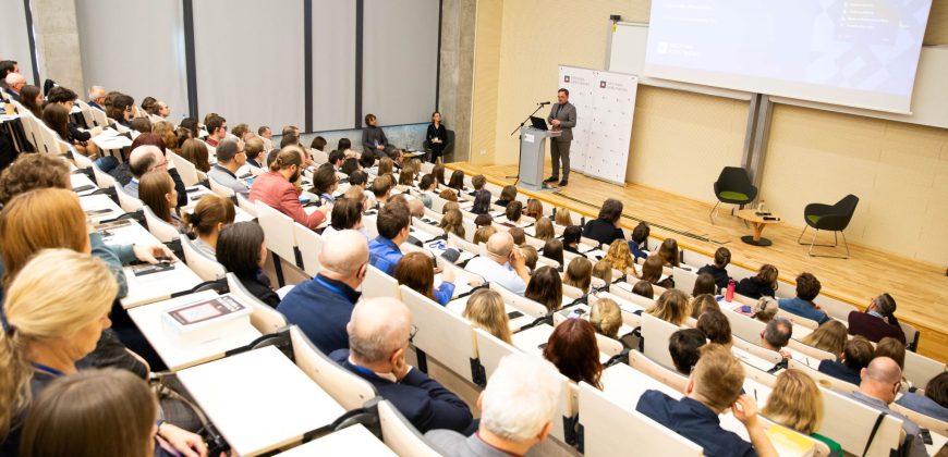 Aula wykładowa. Dyrektor Pietrzyk przemawia do mikrofonu. Za nim rollupy z logo Archiwa Państwowe. Przed nim siedzi publiczność.