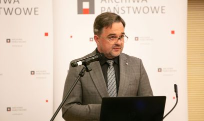 Aula wykładowa. Dyrektor Pietrzyk przemawia do mikrofonu. Za nim rollupy z logo Archiwa Państwowe. Przed nim siedzi publiczność.