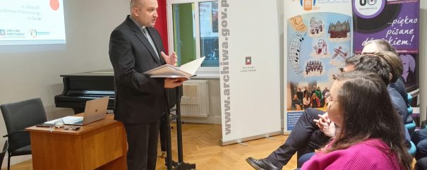 Ks. Poźniak stoi na środku i przemawia do publiczności siedzącej na sali.