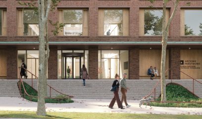 A modern brick building with large windows, a glass entrance, and a wide staircase. Two people walk their dog on the sidewalk in front of the building, while others can be seen inside and on the stairs. Trees and greenery line the path. The sign reads 