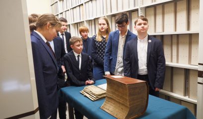 Magazyn. Kilkunastu uczniów stoi dookoła stolika. Na stoliku leży wielka księga archiwalna.