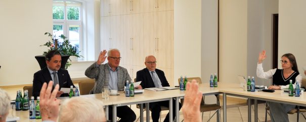 Członkowie rady siedzą za stolikiem i podnoszą ręce w geście głosowania.