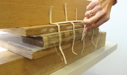 Grzbiet księgi ułożony pomiędzy deskami i połączony paskami materiału. Konserwatorka przesuwa po nim białym narzędziem.